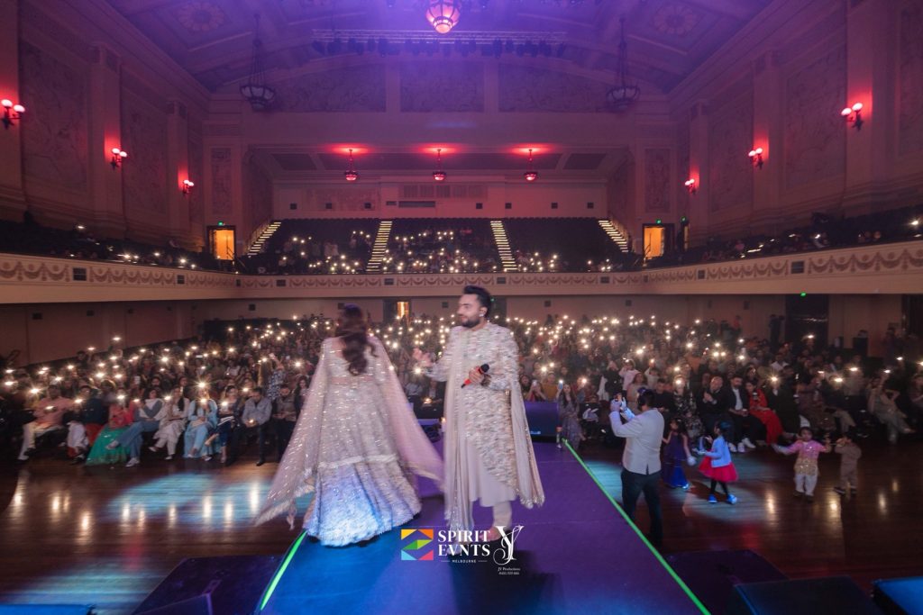 Wide view from the back of Melbourne Town Hall at Bhajan Clubbing, with the audience holding up glowing phone screens towards two performers on the stage runway