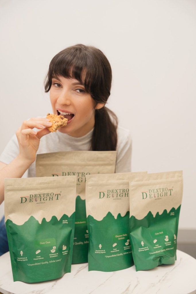 Smiling woman taking a bite of a cookie behind a row of four Dextro Delight Australia product bags on a white marble table
