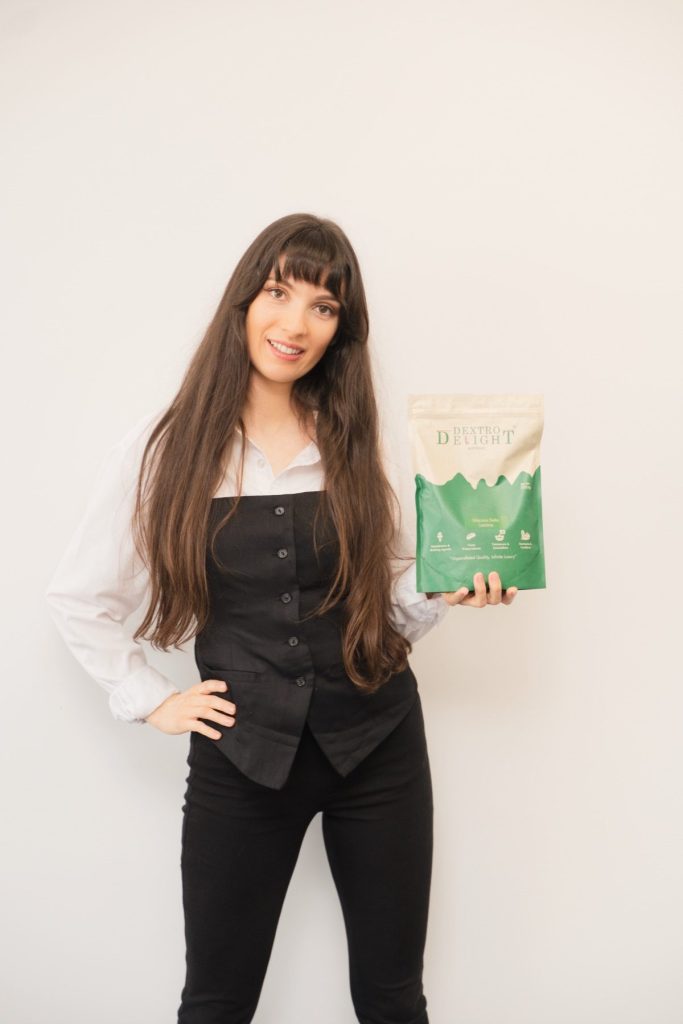 Woman in a white shirt and black vest smiling and holding a Dextro Delight Australia pouch against a white wall