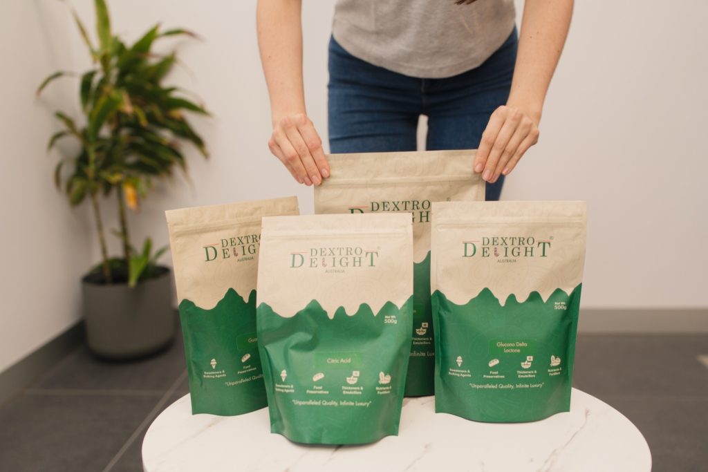 Four Dextro Delight Australia bags arranged on a white marble table with a hand reaching for the centre bag and a green plant behind