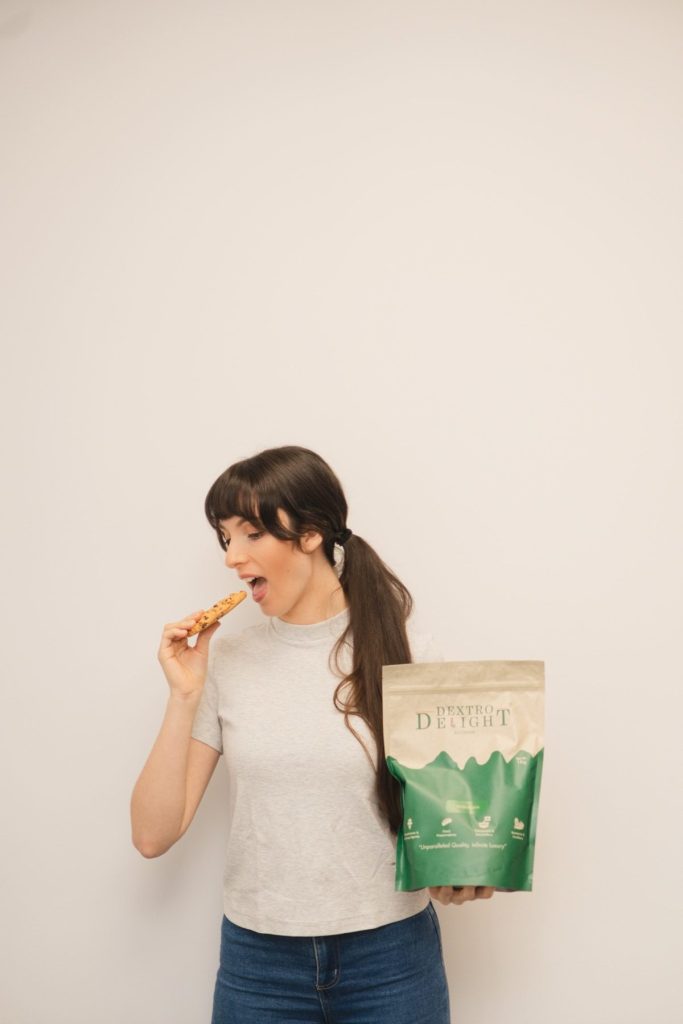 Woman in a grey t-shirt and jeans eating a cookie while holding a Dextro Delight Australia pouch