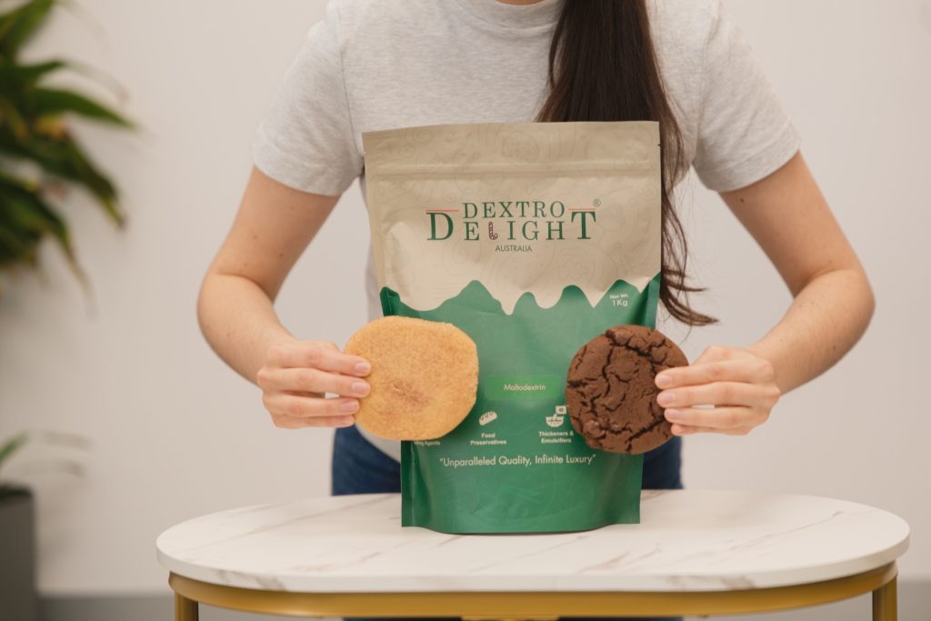 Hands holding a light cookie and a dark chocolate cookie either side of a Dextro Delight Australia bag on a marble table