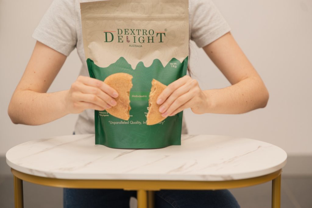 Close-up of hands breaking a large cookie in half over an open Dextro Delight Australia bag with crumbs on the marble table