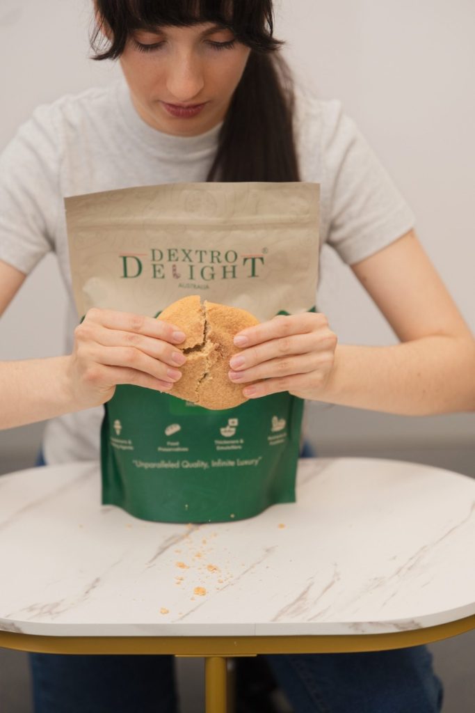 Woman breaking a large cookie over a Dextro Delight Australia bag on a white marble table