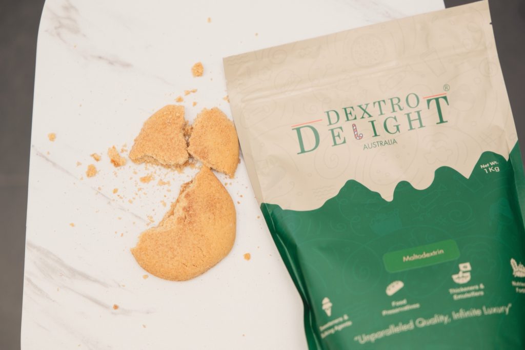 Top-down view of a broken cookie with crumbs next to an open Dextro Delight Australia bag on white marble