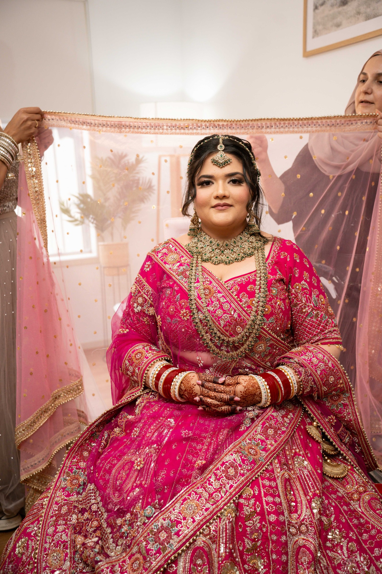 Aman seated in full bridal jewellery while two women hold a sheer pink dupatta over her head