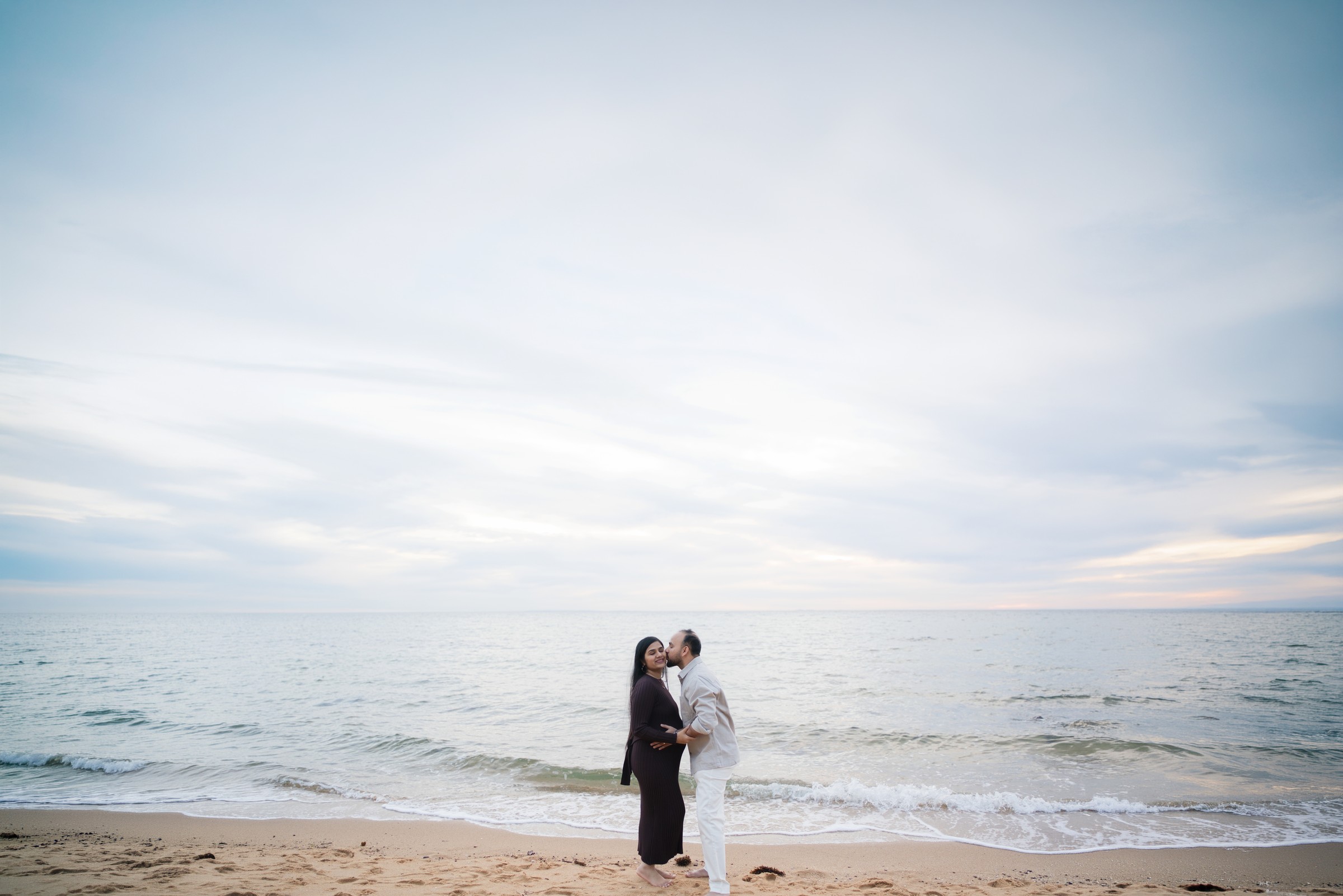 Maulik and Madhvi sharing a kiss on the beach beneath a soft pastel sky