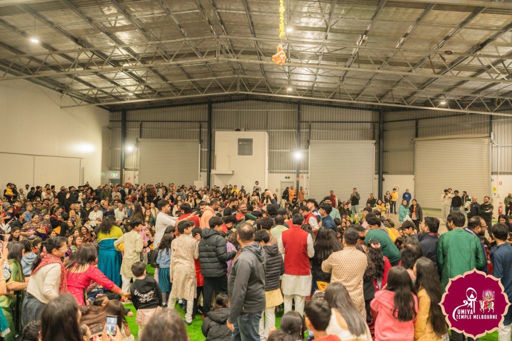 Large crowd of people in colourful traditional clothing gathered in a hall with a high metal ceiling at a Janmashtami celebration in Melbourne