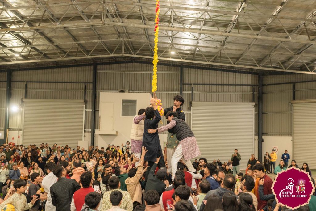 Wide view of the human pyramid reaching for the hanging Dahi Handi pot, surrounded by a large watching crowd at a Janmashtami celebration in Melbourne