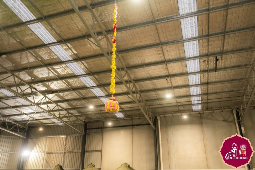 Looking up at the hall ceiling where a decorated Dahi Handi pot hangs from a long yellow flower garland at a Janmashtami celebration in Melbourne