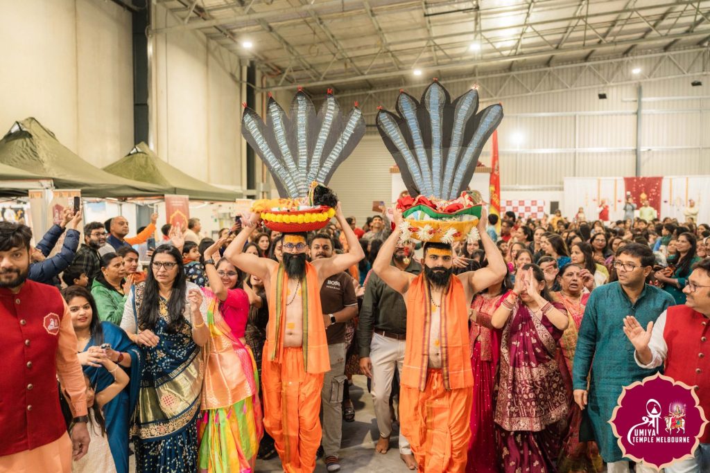 Two men in orange robes carrying large multi-hooded snake headpieces through the crowd in a procession at a Janmashtami celebration in Melbourne